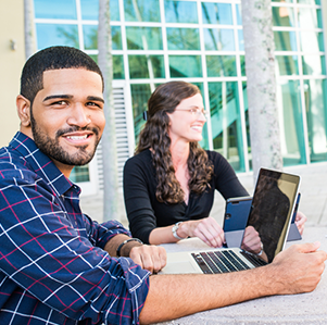 Male and female student sitting at an outdoor table on FAU campus on their laptops. The male is looking at the camera and smiling.